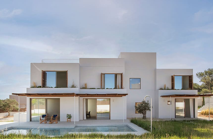 Modern white two-story villa with wooden shutters and a rectangular pool reflecting the sky, bordered by green grass.