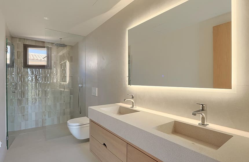 Contemporary bathroom with a light-toned wooden vanity, integrated stone double sink, large backlit mirror, and glass-enclosed shower.