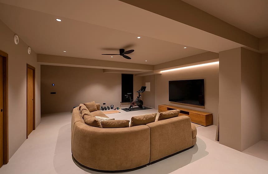 Contemporary media room featuring a large curved beige sofa, wall-mounted screen, wooden console, and stationary bike.
