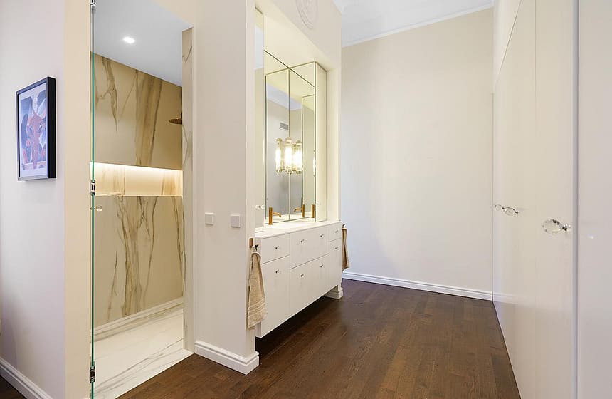 A modern bathroom featuring a walk-in shower with marble-veined walls, a white vanity with gold hardware, and dark wood flooring.