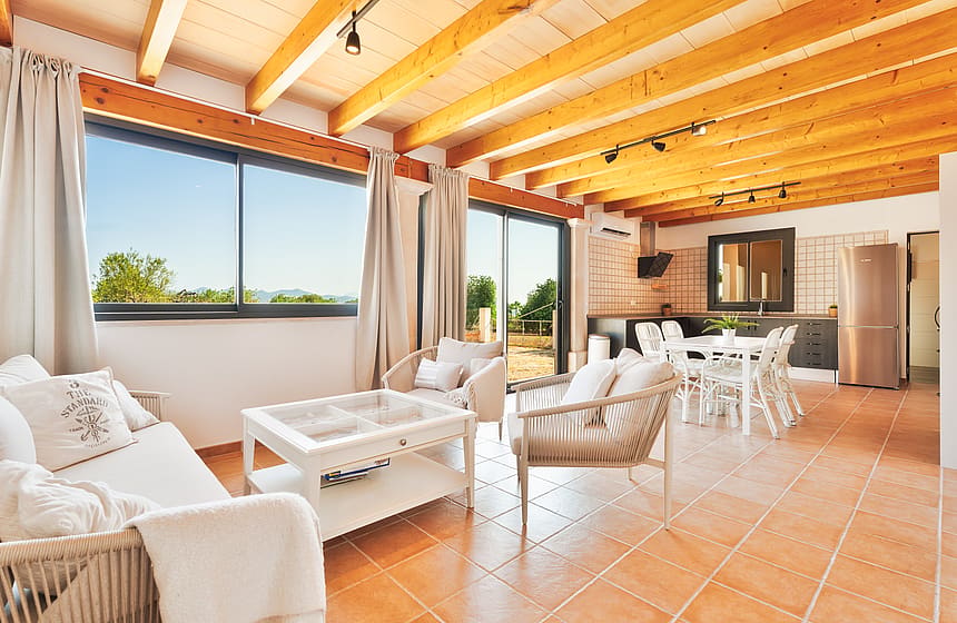 Salón y comedor abierto con vigas de madera vistas, suelos de baldosas y grandes ventanales.