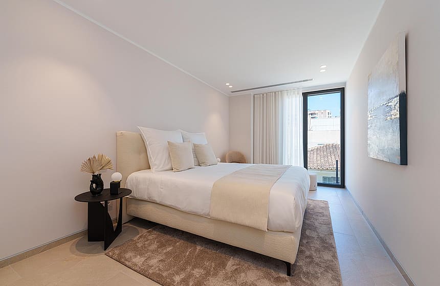 Dormitorio minimalista con cama en tonos neutros, mesita oscura y puerta de balcón con luz natural.
