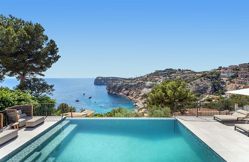 An infinity edge swimming pool with turquoise mosaic tiles overlooking a sea bay with rocky cliffs and pine trees.