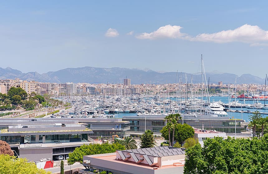 Vista elevada del puerto con yates, el perfil urbano y las montañas costeras bajo un cielo azul.