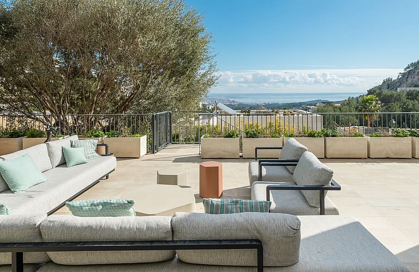 Outdoor terrace lounge with stone flooring, metal and fabric furniture, olive tree, and views of the Mediterranean coastline.