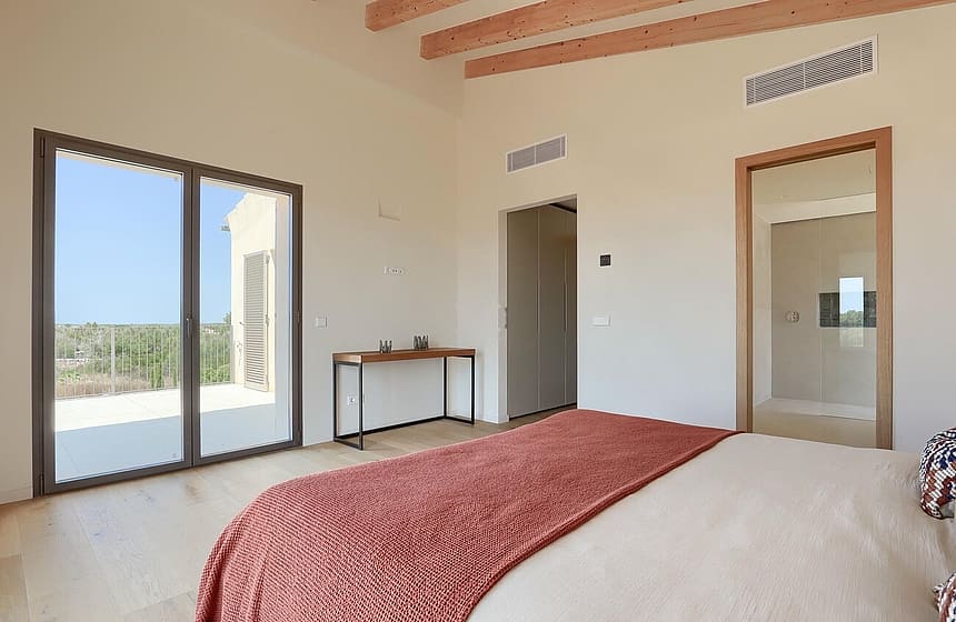 A bedroom with light wood floors, a rust-colored bed throw, and exposed timber ceiling beams, featuring sliding glass doors.