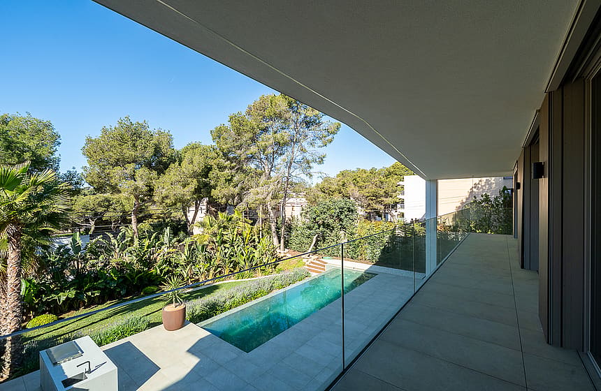 Covered concrete terrace with glass railing looking down onto a narrow rectangular lap pool surrounded by pine trees.