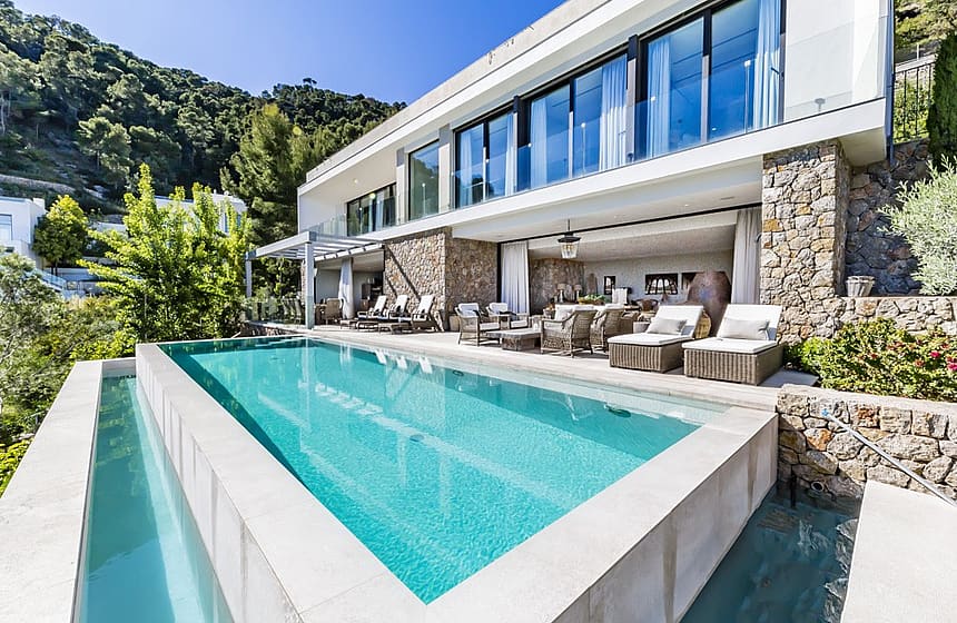 Exteriör av en modern vit villa med stenfasad, infinity-pool med turkost vatten och en möblerad terrass.