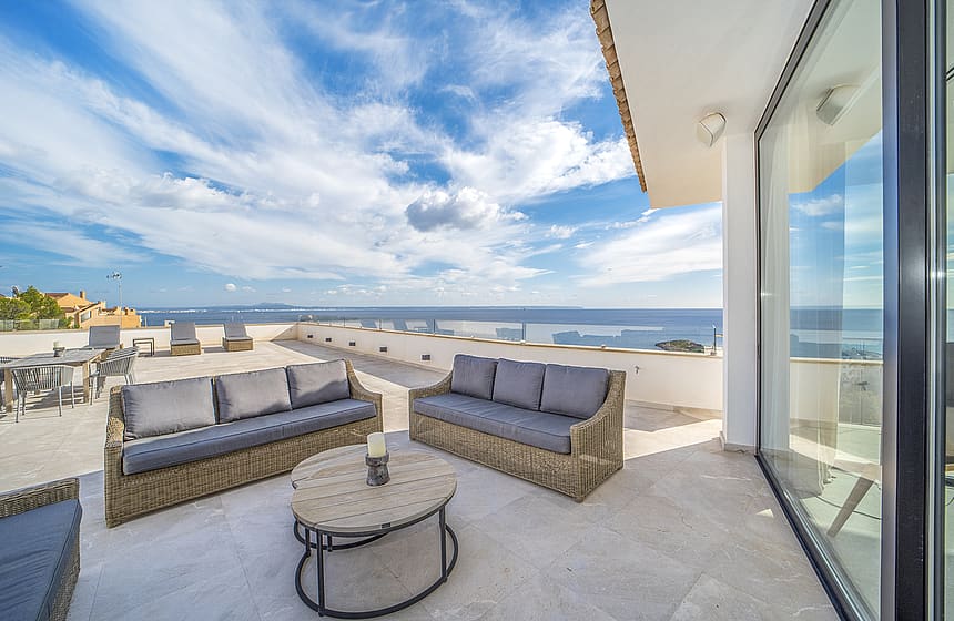 A wide outdoor terrace featuring woven lounge furniture, stone flooring, and expansive views of the sea under a blue sky.