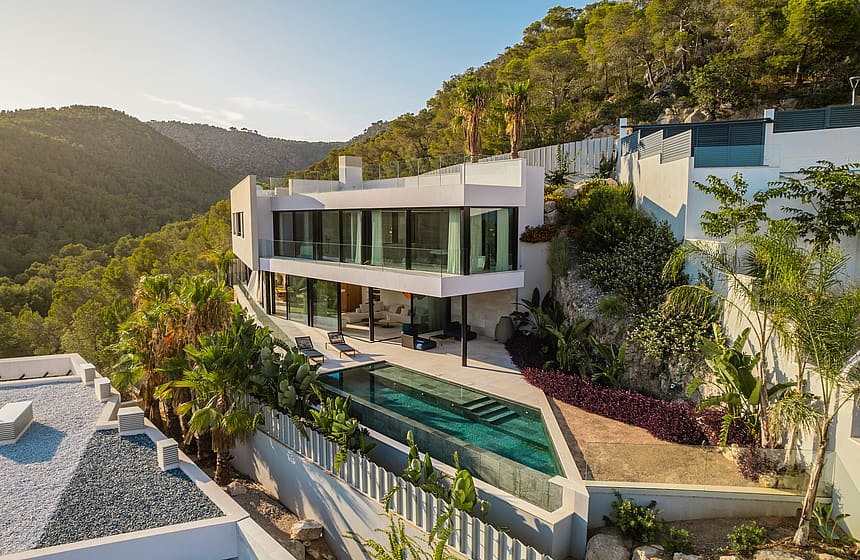 A contemporary white villa built into a hillside with floor-to-ceiling glass walls, a tiered terrace, and a private pool.