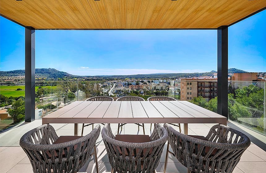 A covered terrace with a wood-paneled ceiling, a long dining table, and dark woven chairs overlooking a landscape of mountains.
