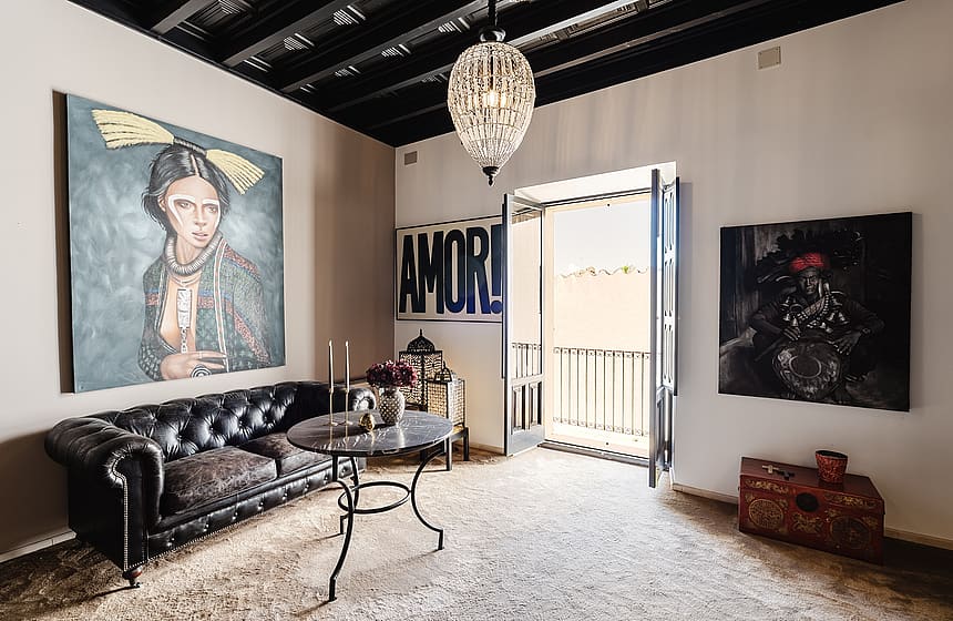 A living room in a Sencelles townhouse featuring a black leather tufted sofa, a glass-topped table, and large-scale artwork on white walls with a black exposed beam ceiling.