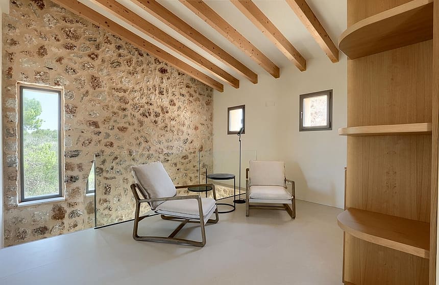 A living space featuring a textured dry-stone wall, exposed wooden ceiling beams, and two neutral-toned armchairs on a smooth floor.