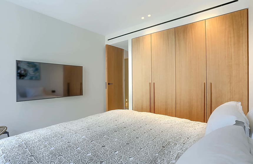 A modern bedroom with white walls, a large bed with patterned bedding, and a wall of floor-to-ceiling light oak wardrobes.