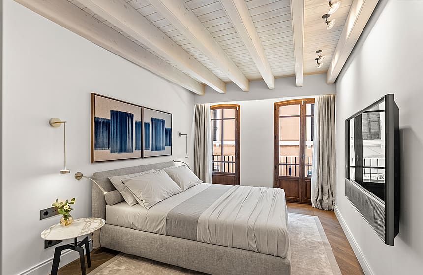 A bedroom featuring exposed timber ceiling beams, herringbone oak floors, and two arched glass doors opening to balconies.