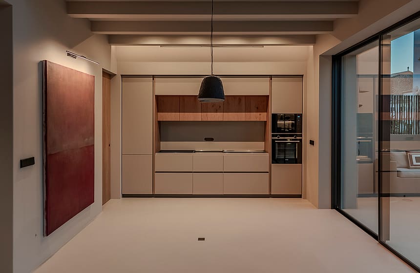 A modern kitchen featuring handleless beige cabinetry, exposed ceiling beams, and large sliding doors opening to a terrace.