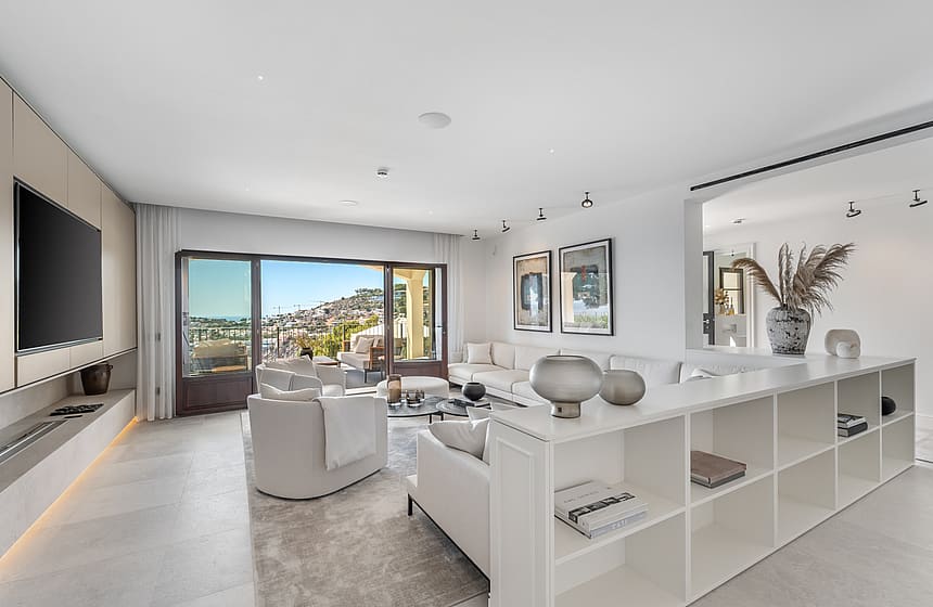 Bright living room featuring white sofas, stone floors, and large sliding doors opening to a terrace with coastal views.