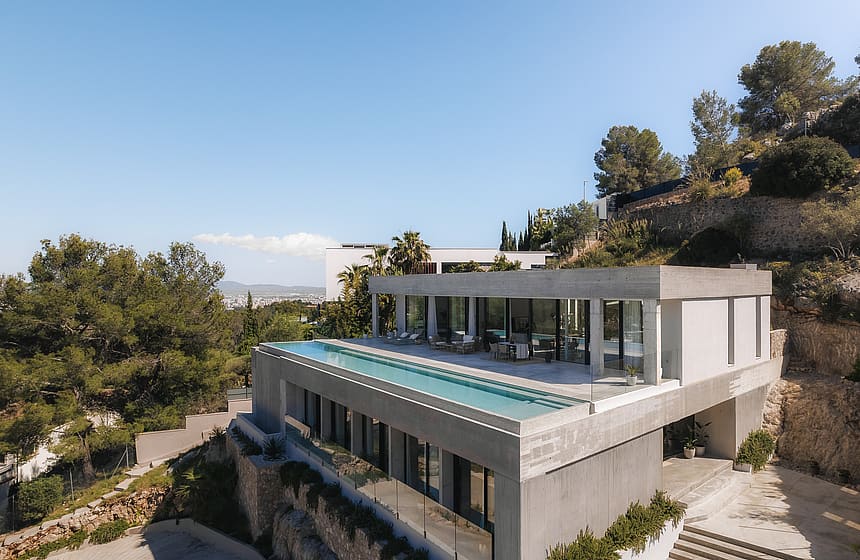 Elevated view of a contemporary concrete villa featuring a rooftop infinity pool and floor-to-ceiling glass walls.