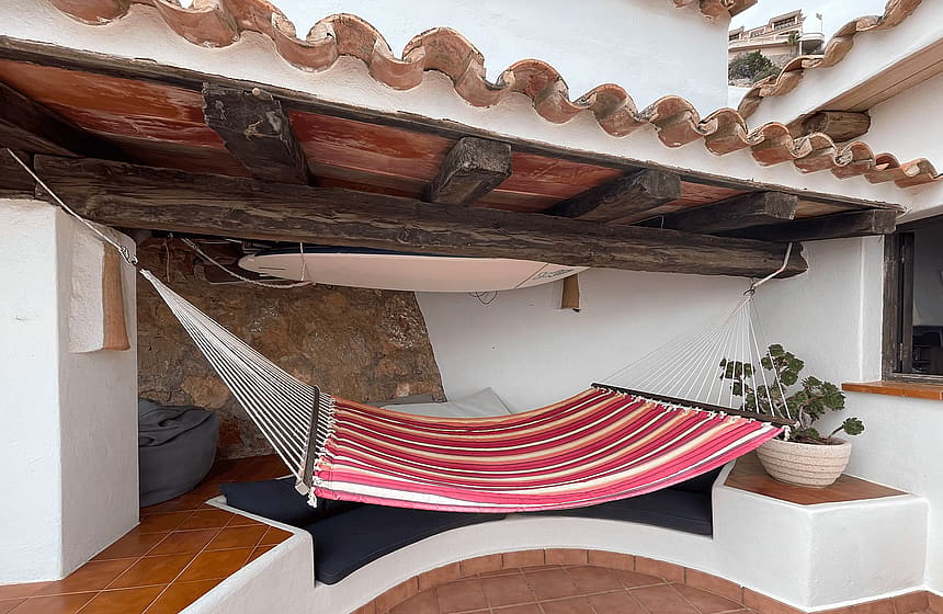 A covered terrace nook featuring a striped fabric hammock strung between white walls under a rustic wooden beam roof.