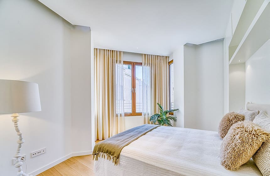A bright bedroom with hardwood floors, a white bed with textured pillows, and beige floor-to-ceiling curtains.