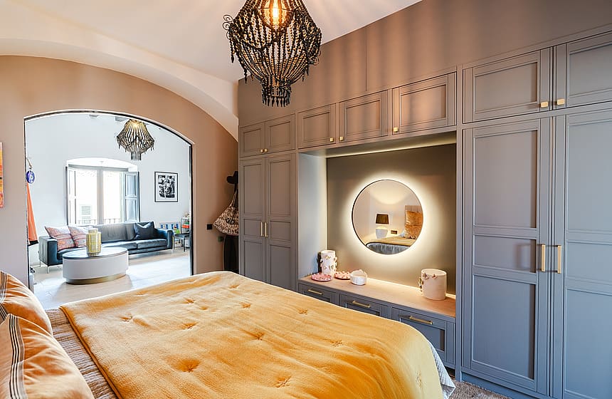A master bedroom featuring a large bed with a golden throw, grey paneled wardrobes, a circular backlit mirror, and an arched doorway leading to a sitting area.