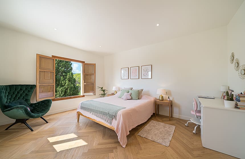 A bright bedroom featuring light oak herringbone parquet, a green velvet armchair, and wooden shutters opening to trees.