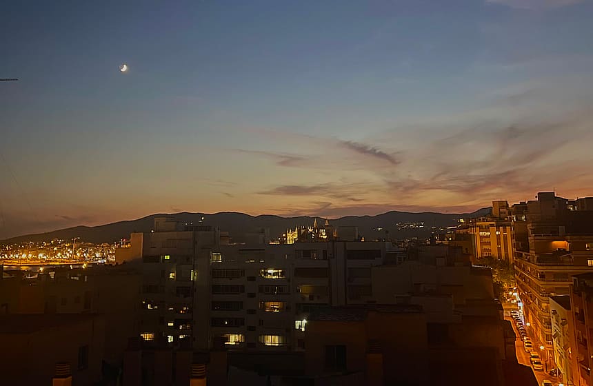 A dusk view over a city with warm street lights and building windows glowing under a twilight sky with a visible moon.
