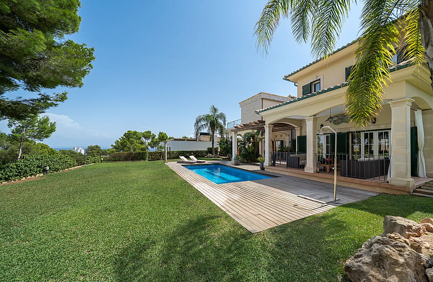 Exterior view of a villa with a rectangular swimming pool, manicured lawn, sandstone columns, and a shaded terrace.