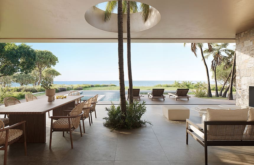 A wide outdoor covered terrace featuring a dining area, lounge seating, and an open circular skylight with trees, facing the sea.