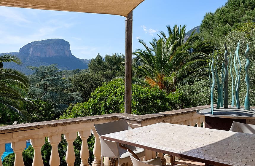 An outdoor terrace with a stone balustrade, a stone dining table, and a shaded seating area overlooking Alaro mountain peaks.