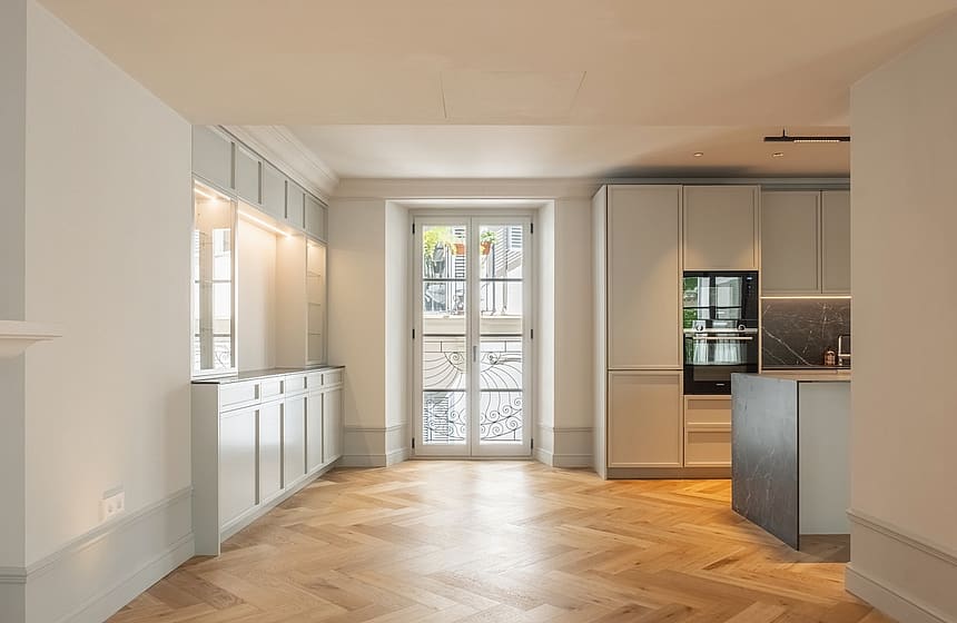 Interior view of a renovated apartment featuring light oak herringbone flooring, contemporary kitchen cabinetry, and French doors.
