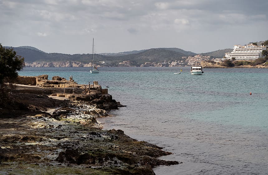 Rocky Mediterranean shoreline with turquoise water, a distant sailboat, and coastal hills under an overcast sky.