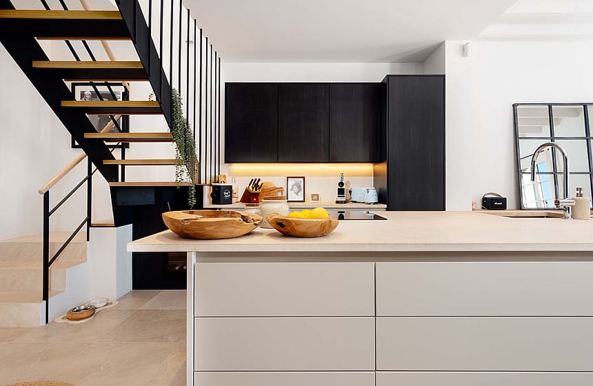 Modern kitchen with matte black cabinetry, stone-topped island, and an open-tread metal and wood staircase in the foreground.