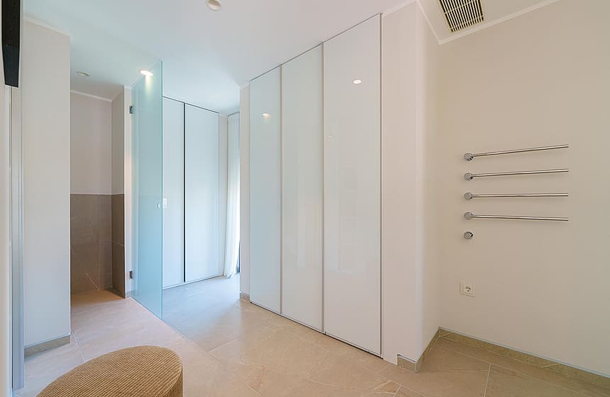 Vestidor luminoso con armarios blancos de suelo a techo, partición de cristal, taburete y toalleros.