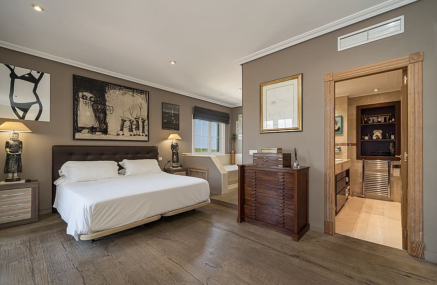 A master bedroom featuring a large bed, dark wooden dresser, and an open doorway leading into a tiled en-suite bathroom.