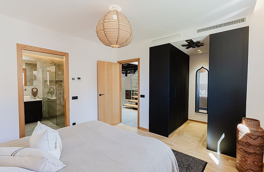 A master bedroom featuring light wood flooring, a beige bedspread, a black wardrobe unit, and an open doorway to a marble bathroom.