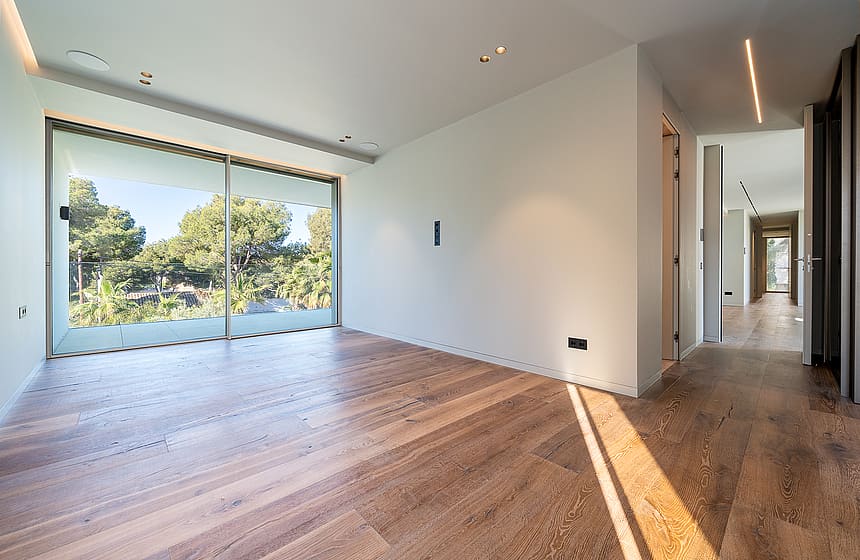 Empty modern bedroom with wide oak floorboards, white walls, and a large sliding glass door looking out toward green trees.