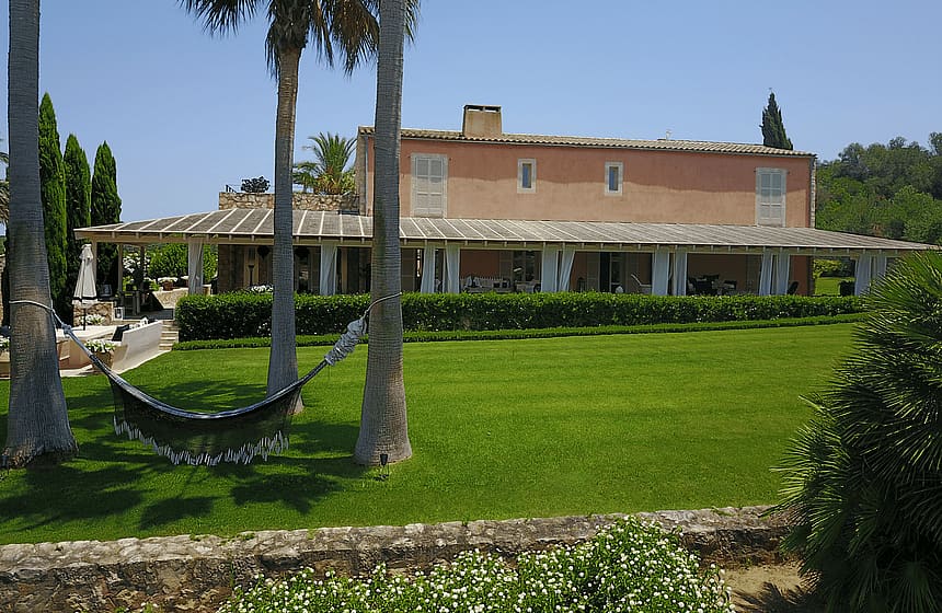 Exterior view of a traditional terracotta-toned finca with a long covered porch, palm trees, and a hammock on a green lawn.