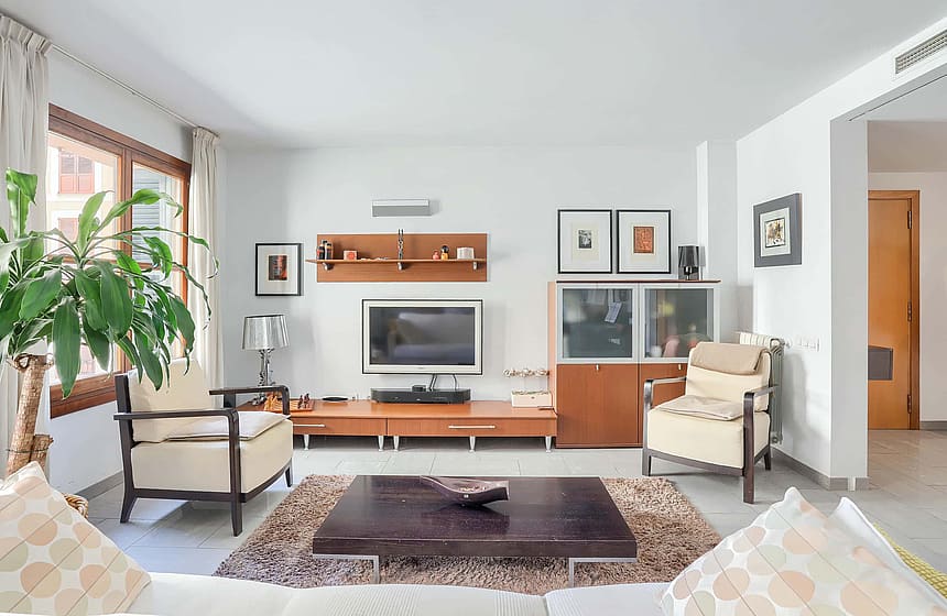Living room featuring cream armchairs, a low dark wood coffee table, and a wall-mounted television unit on white walls.