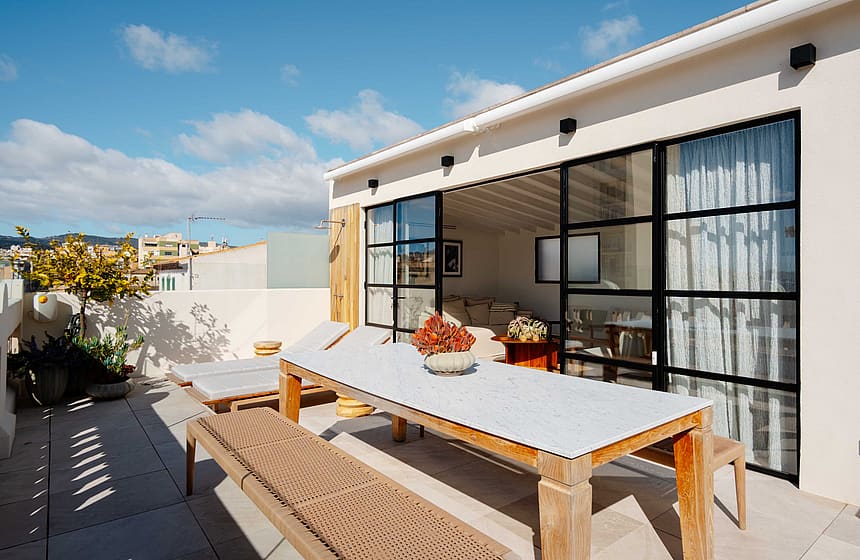 A rooftop terrace featuring a wooden dining table and benches, connected to an indoor living area by black-framed glass doors.
