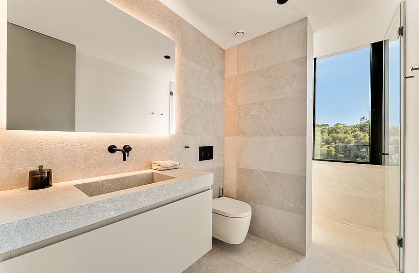 A minimalist bathroom featuring large-format beige tiles, a wall-hung toilet, a floating vanity with an integrated mirror, and a glass-enclosed shower area with views of greenery.