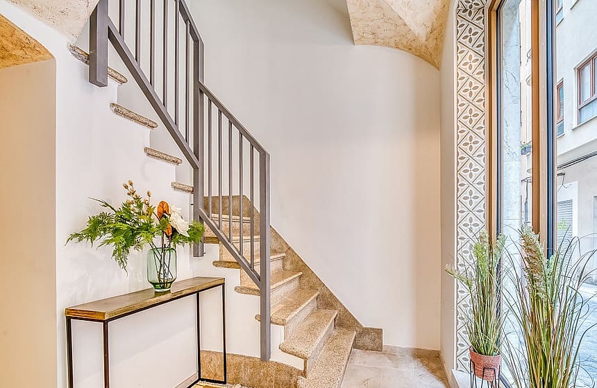 Interior entrance hall featuring a light stone staircase with a dark metal railing, a slim console table, and patterned tiles.