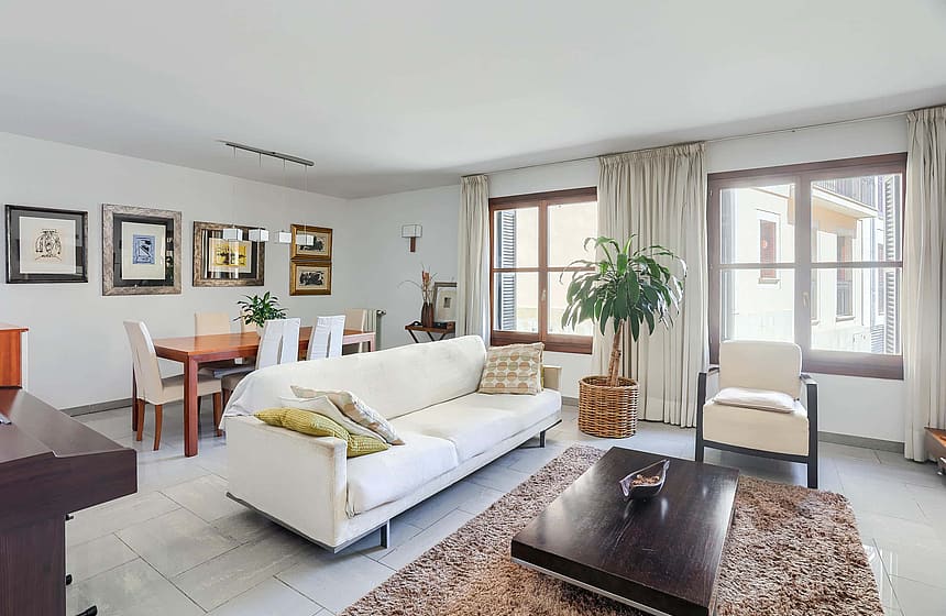 A bright living room with a white sofa, dark wooden coffee table, and a dining table for six, featuring neutral decor.