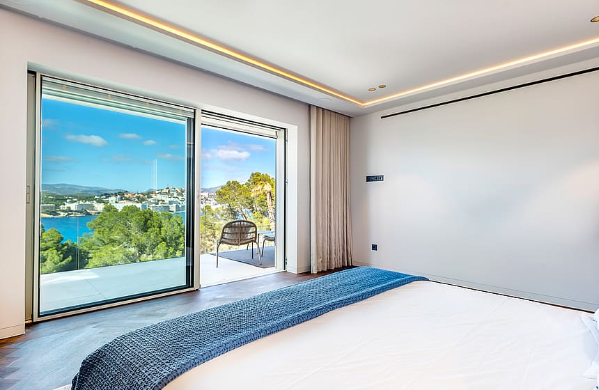 A modern bedroom with floor-to-ceiling sliding glass doors opening onto a terrace overlooking a turquoise coastal bay.