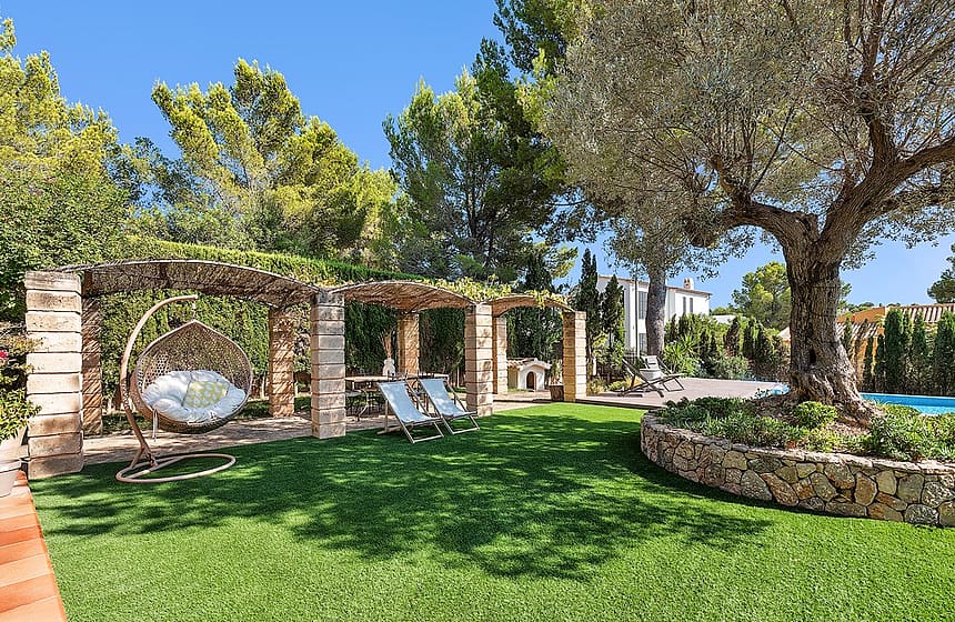 A green manicured lawn with a stone pergola, hanging wicker swing chair, and an mature olive tree in a sunny garden.