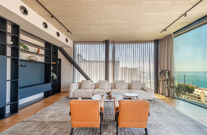 Contemporary living room with a large cream sofa, two orange leather armchairs, and floor-to-ceiling glass overlooking the sea.