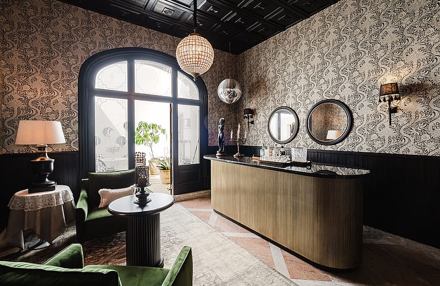 A formal bar area featuring patterned wallpaper, black wainscoting, a gold-toned bar counter, and velvet armchairs near a large arched window.