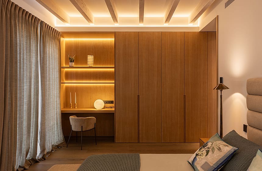 Bedroom featuring custom floor-to-ceiling wooden wardrobe, built-in shelving desk area, and neutral fabric curtains.
