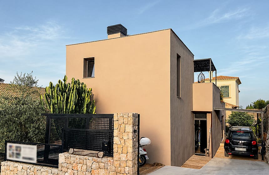 Exterior view of a beige concrete villa in Genova featuring a stone perimeter wall, black metal gate, and a private driveway.