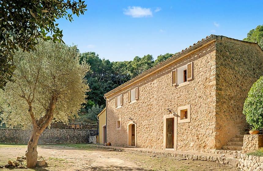 Exterior view of a traditional stone finca with timber shutters, surrounded by olive trees and pine forest under a blue sky.